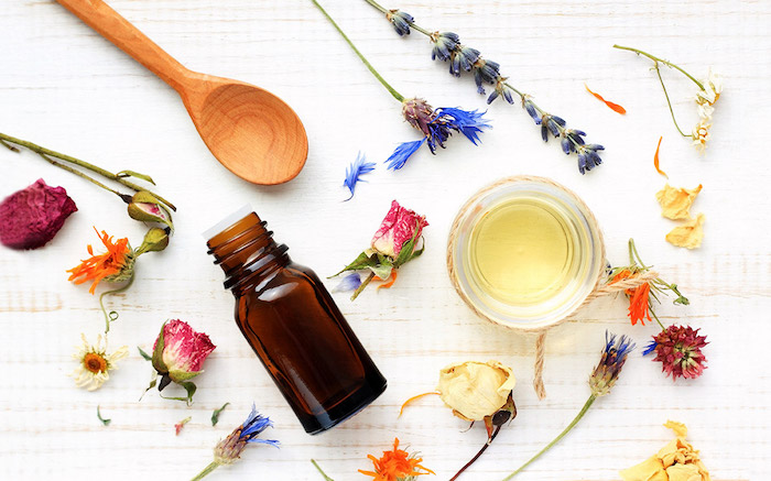 des herbes et des fleurs sur une nappe blnache a coté d une cuillère en bois et un pot d huile essentiel