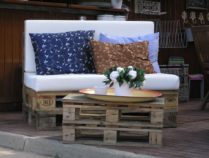 canape exterieur palette qvec des coussin blu et beige petite table en bois avec des fleurs blanches dessus