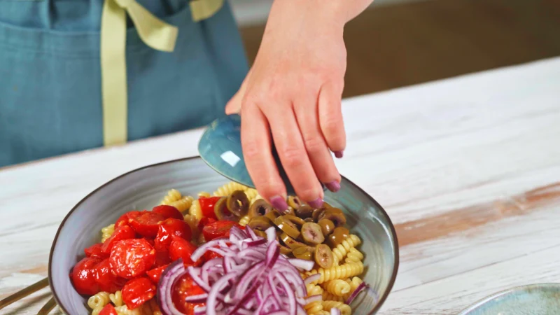 une main qui verse des produits dans un bol de pates tomates et olives