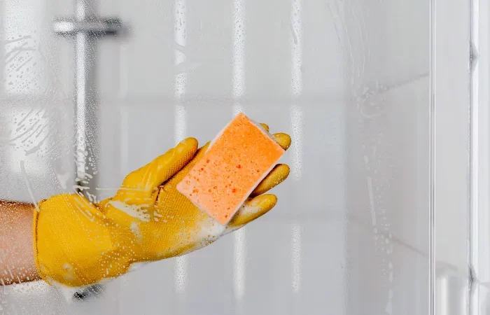 une main avec un gant jaune et eponde orange nettoie le plexiglas de la douche