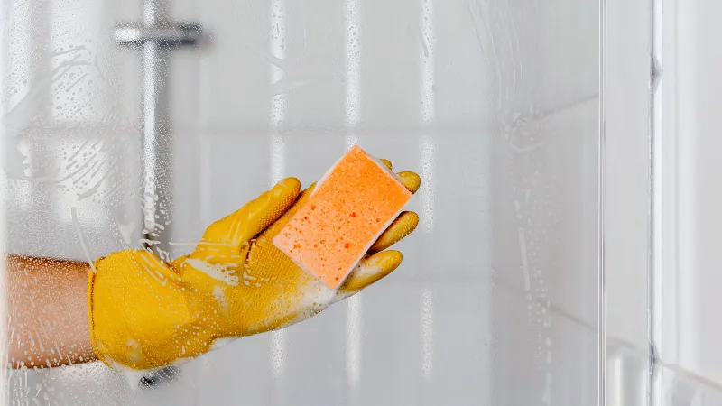 une main avec un gant jaune et eponde orange nettoie le plexiglas de la douche