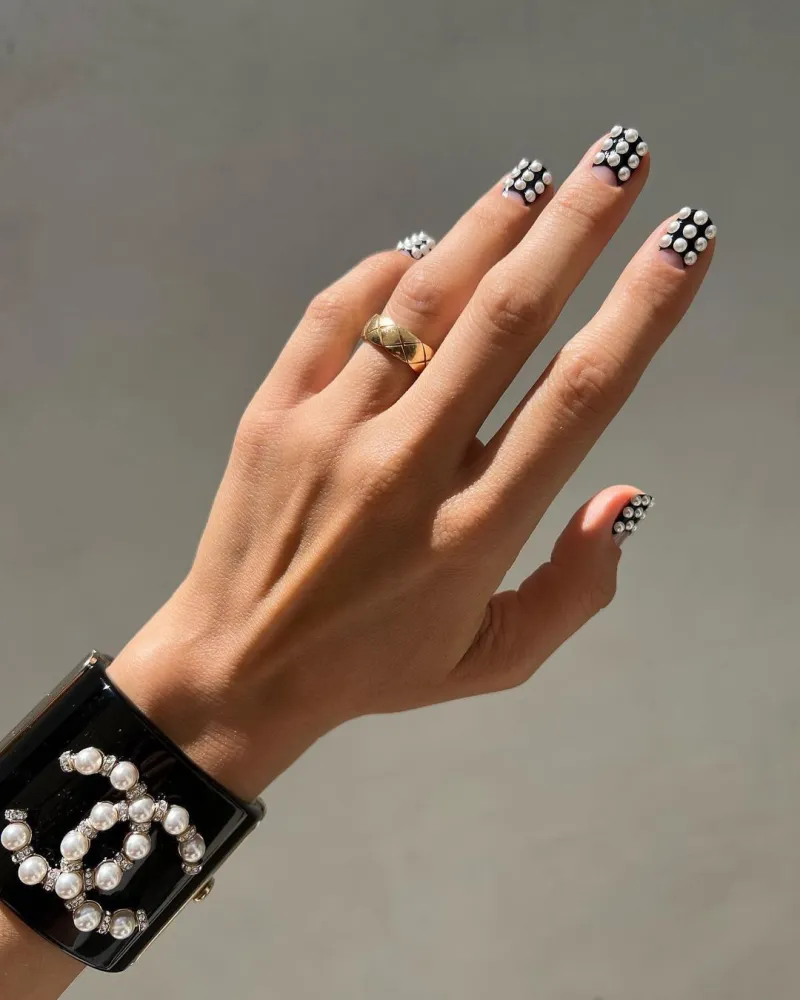 une main avec une bague et des ongles vernis en demie lune noir avec des perles et un bracelet chanel sur le poignet sur fomd gris