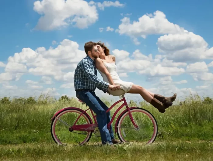 un couple heureux sur un velo dans un champs sur fond de ciel bleu avec quelques nuages blanc