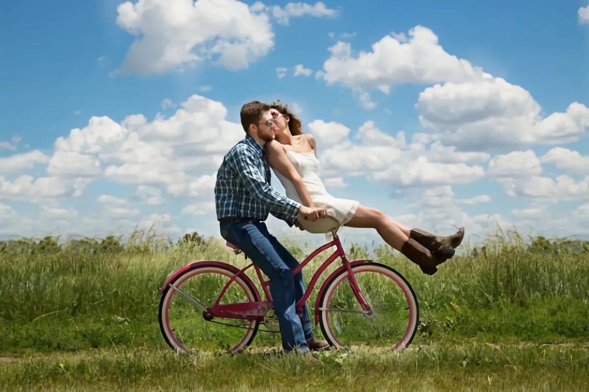 un couple heureux sur un velo dans un champs sur fond de ciel bleu avec quelques nuages blanc