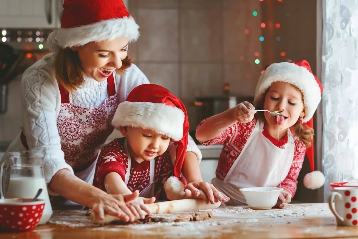 une famille qui fabrique des biscuits de noel avec un adulte et deux enfants portants tous des chapeaux de noel