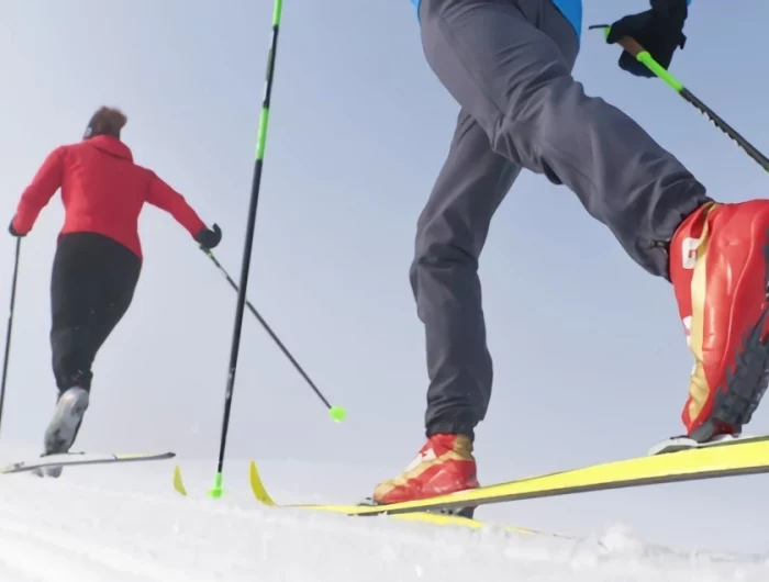 une personne de pleine taille qui fait du ski de fond avec une deuxieme en premier plan avec les jambes visibles uniquement