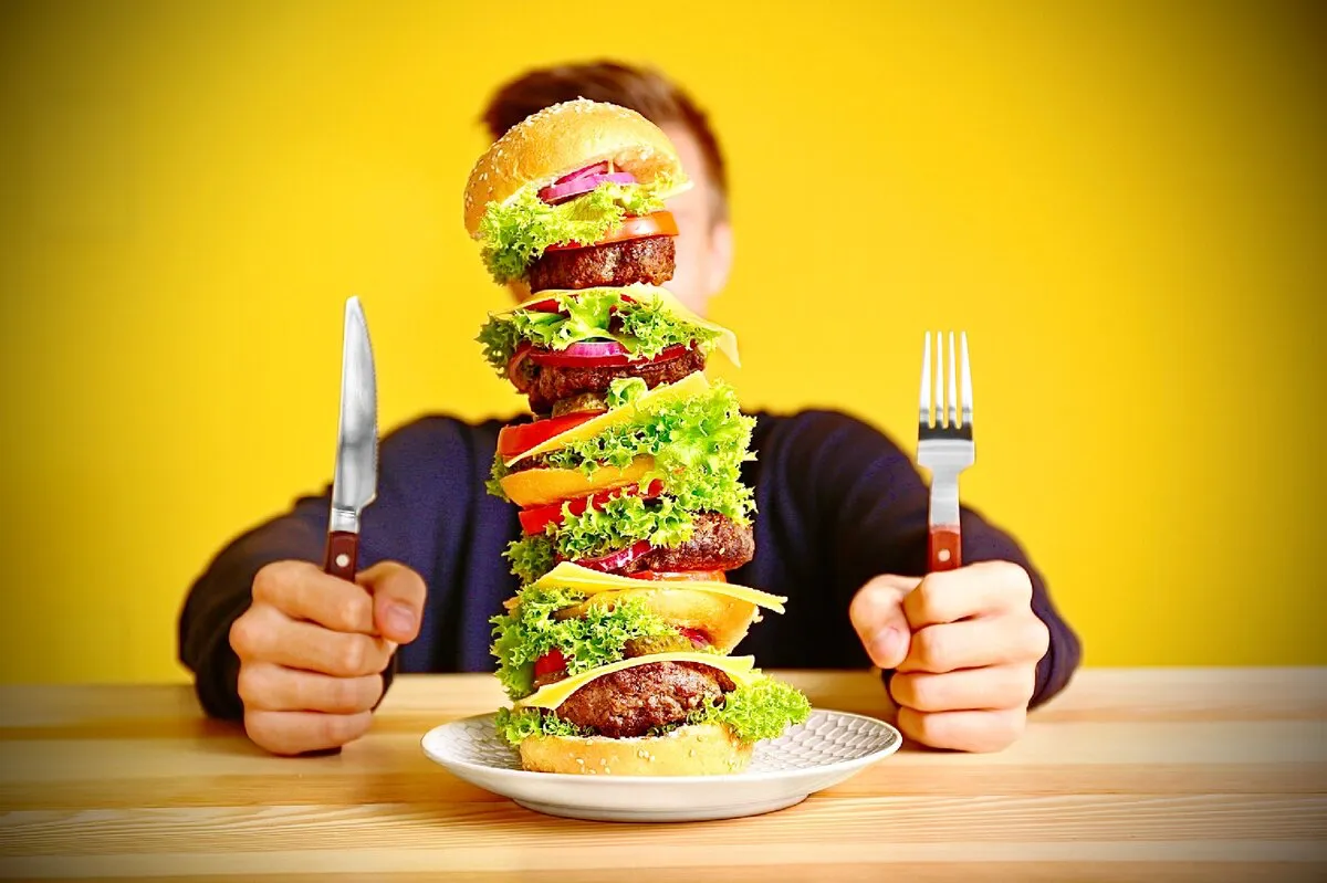 un mega burger qui cache le visage de celui qui va le manger qui tient une fourchette et un couteau sur une table en bois et un mur jaune