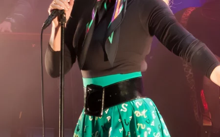arielle dombasle sur scene au micro habillee avec une mini jupe verte avec un imprime de lettres noires et blanches portant une ceinture noire avec un haut noir