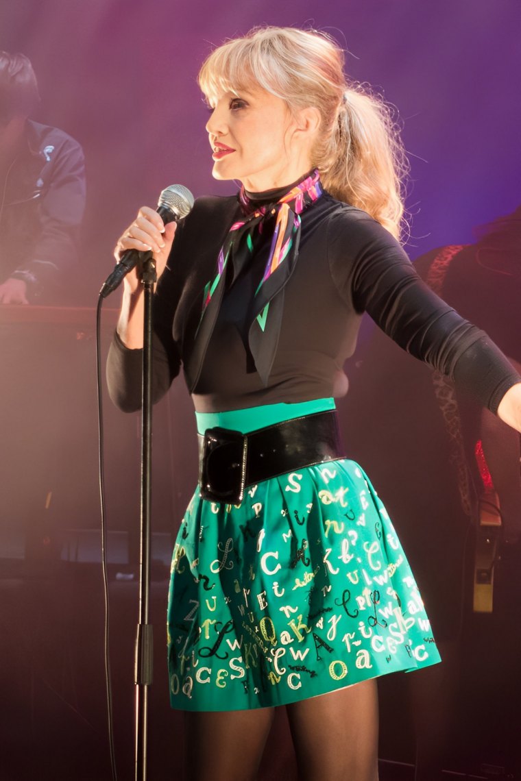 arielle dombasle sur scene au micro habillee avec une mini jupe verte avec un imprime de lettres noires et blanches portant une ceinture noire avec un haut noir