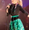 arielle dombasle sur scene au micro habillee avec une mini jupe verte avec un imprime de lettres noires et blanches portant une ceinture noire avec un haut noir