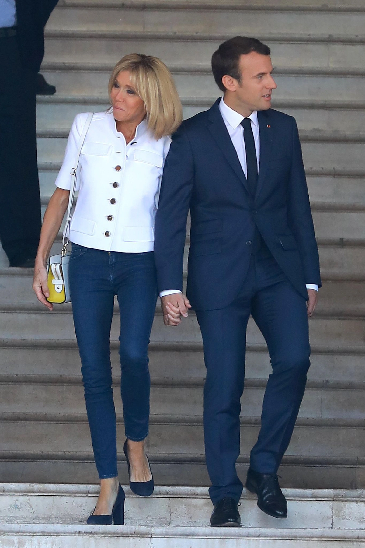 brigitte macron main dans la main avec son mari mais regardant dans des dire rions opposees habillee en jeans avec des talons et veste blanche courte