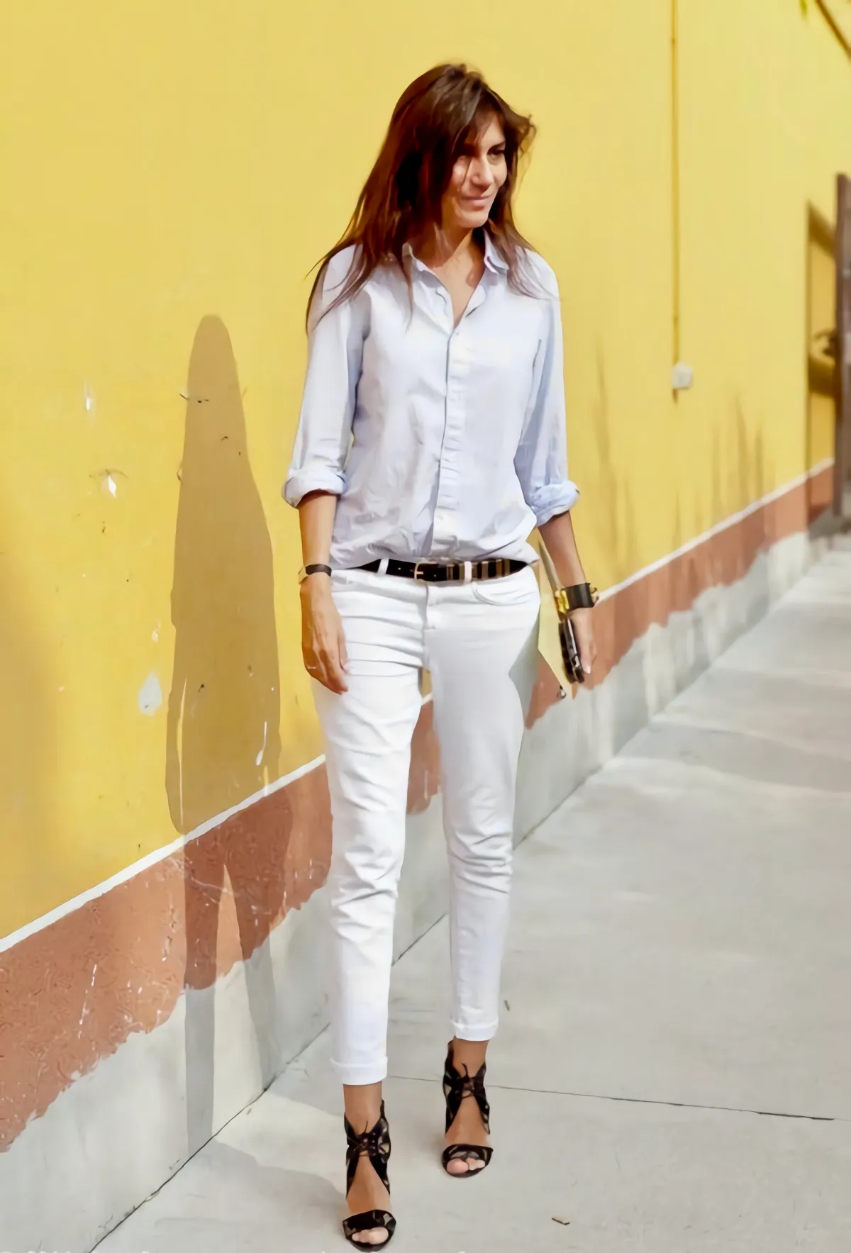 emmanuelle alt en jeans blanc ceinture noir escarpins noirs et che;ise a manches retroussees sur fond un mur jaune