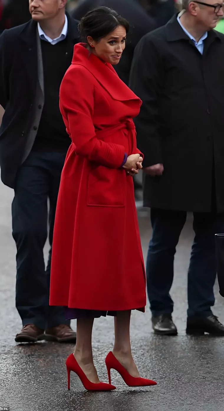 meghan fait une grimace habillee en long manteau rouge et de chaussures beiges a talons rouges