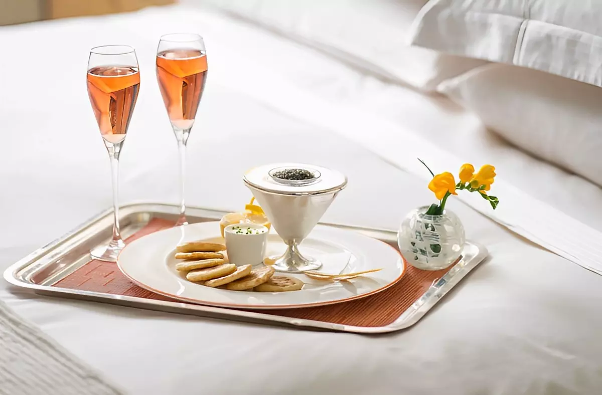 plateau sur un lit aux draps blancs avec une assiette remplie de blinis caviar noir sauce et citron a cote de deux verres de champagne