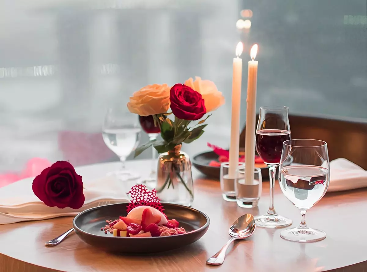 table dressee avec des bougies un petit bouquets de roses des verres remplies de vin rouge et deux assiettes avec un dessert de fruits rouges et des couverts