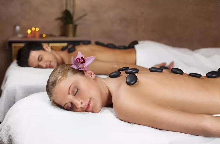 un homme et une femme avec une fleure dans les cheveux sont allonges sur le ventre sur deux tables de massage avec des pierres sur le dos
