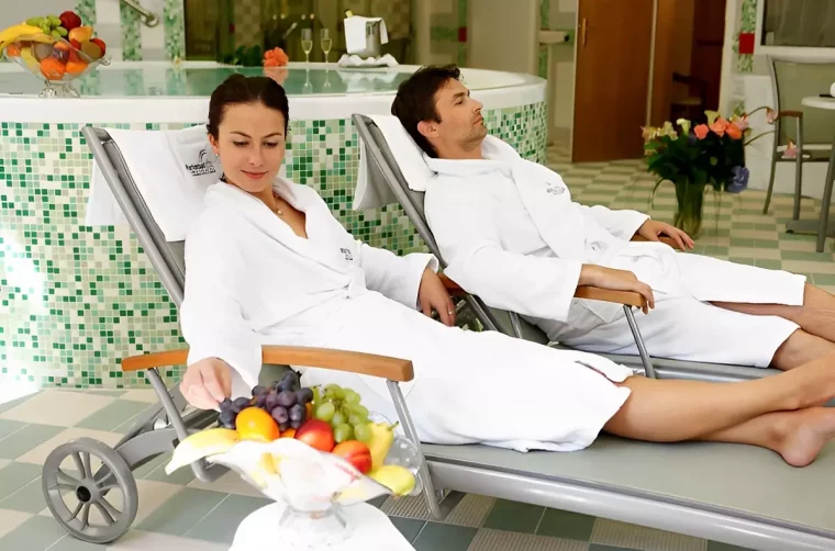un homme et une femme souriante allonges sur deux chaiselongues a cote du jacouzzi et des fruits en premier plan