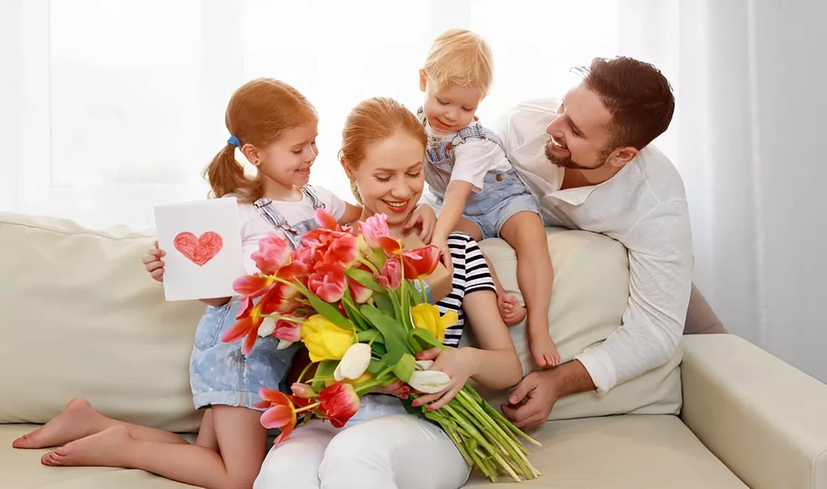 une famille avec deux enfants et un homme souriant regardant sa femme assise sur un canape beige tient un bouquet de fleurs