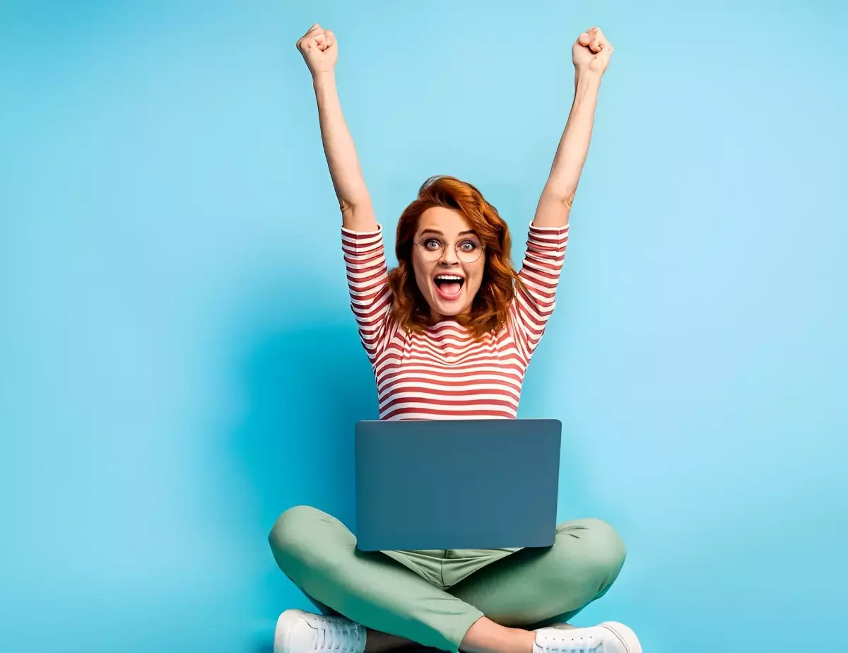 une femme sur fond bleu qui montre de l enthousiasme assise avec les jambes croisees et les bras souleves avec un ordinateur portable devant elle