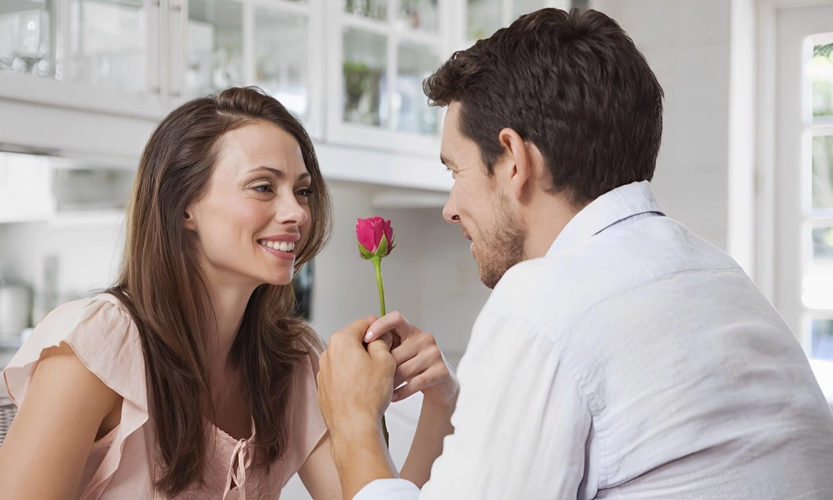 un homme en chemise blanche assis de dos offre une rose rouge a une femme qui le regarde dans les yeux et lui sourit