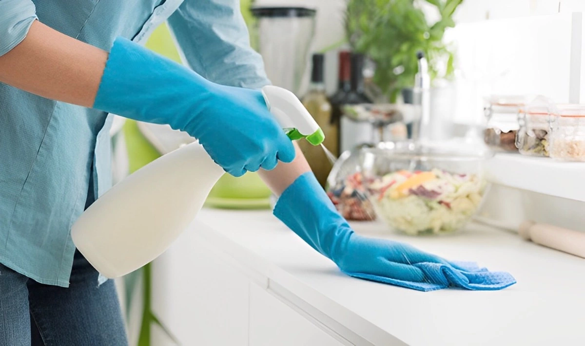une personne en chemise bleue et des gants de cuisine bleus nettoie sa cuisine avec un produit dans une main et une eponge dans l autre