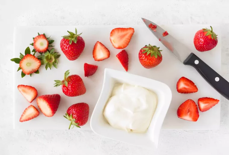 fraises entieres et fraises decoupees avec un couteau et un petit bol blanc avec un produit laitier sur une planche blanche et fond blanc