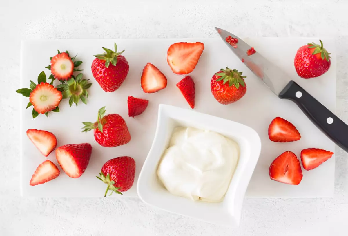 fraises entieres et fraises decoupees avec un couteau et un petit bol blanc avec un produit laitier sur une planche blanche et fond blanc