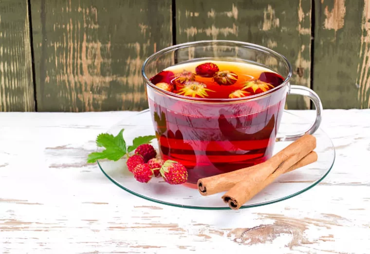 infusion dans une tasse en verre avec des petites fraises qui flottent et deux battons de cannelle sur la sous tasse sur fond en bois bi couleur