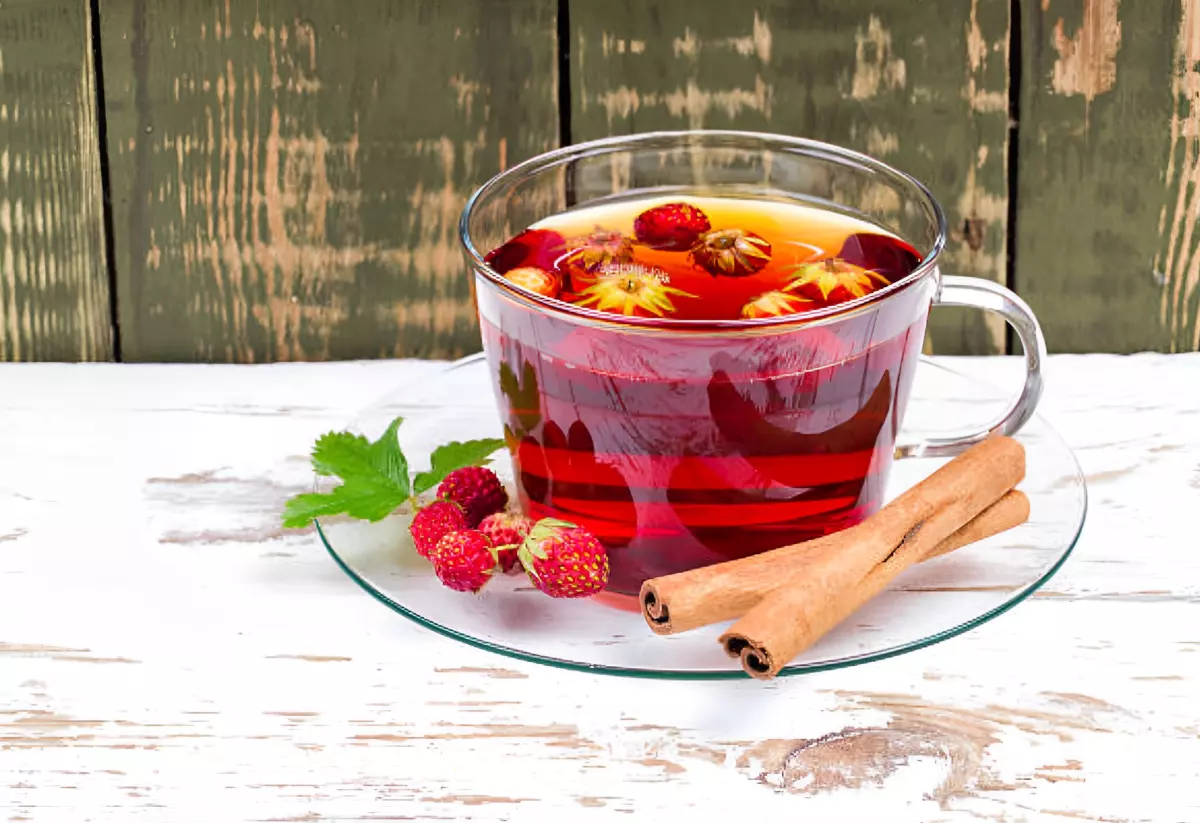 infusion dans une tasse en verre avec des petites fraises qui flottent et deux battons de cannelle sur la sous tasse sur fond en bois bi couleur