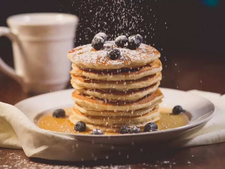 un tas de pancakes sur un lit de sauce avec des myrtilles par dessus et autour dans une assiette blanche et une tasse blanche derriere