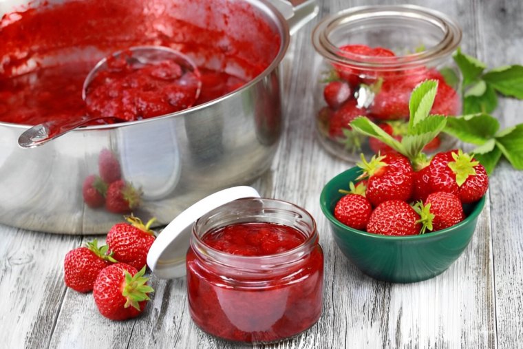 une louche dans une casserole avec de la confiture de fraises avec des pots pleines de la confiture et des fraises fraiches sur un plancher en bois claire