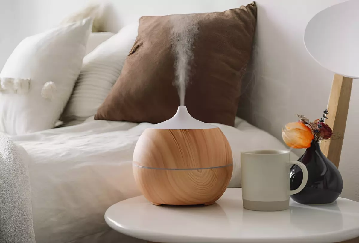 diffuseur d huiles essentielles pose a cote d une tasse sur une table basse blanche devant un lit avec des coussins blancs et un marron