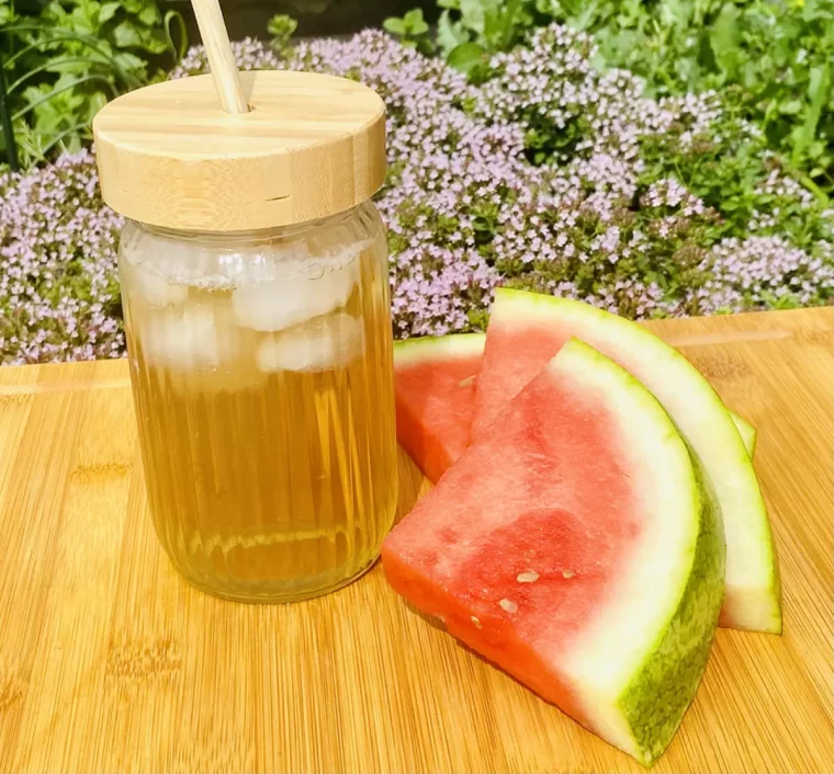 infusion de pasteque dans un verre avec un couvercle des glacons et une paille a cote de deux tranches de pasteque sur une surface en bois sur fond de fleurs