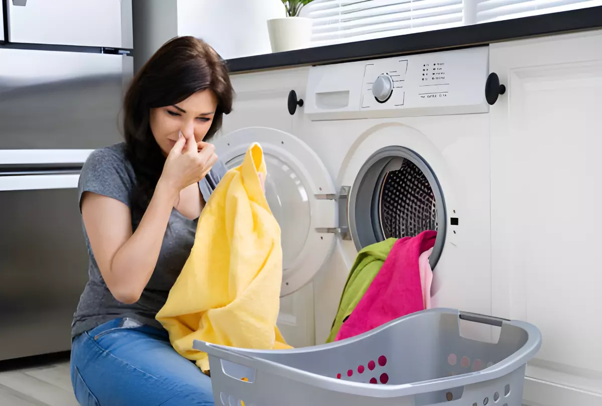 femme assise au sol devant une machine a laver et un panier a linge tient une serviette jaune dans la main gauche et bouche son nez avec la main droite