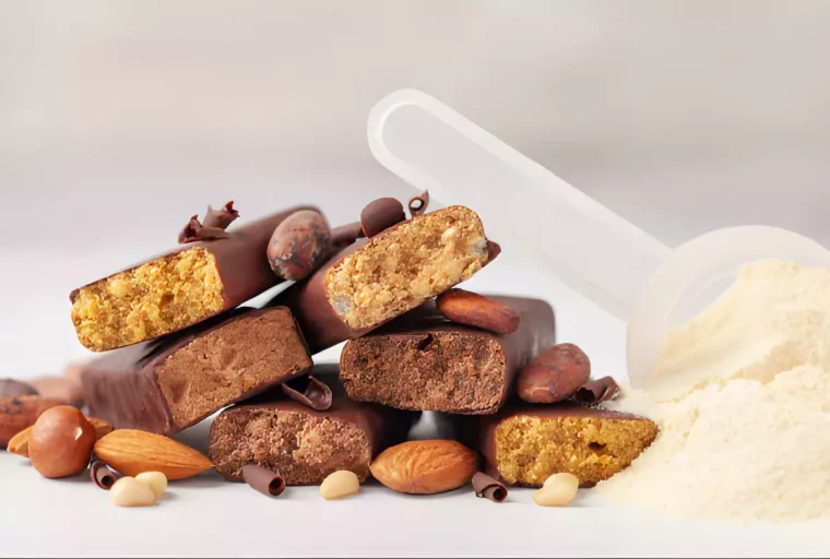 un tas de bars proteinees avec des sciures de chocolat quelques amandes noisette et autres noix autour et de la poudre de protéines a droite