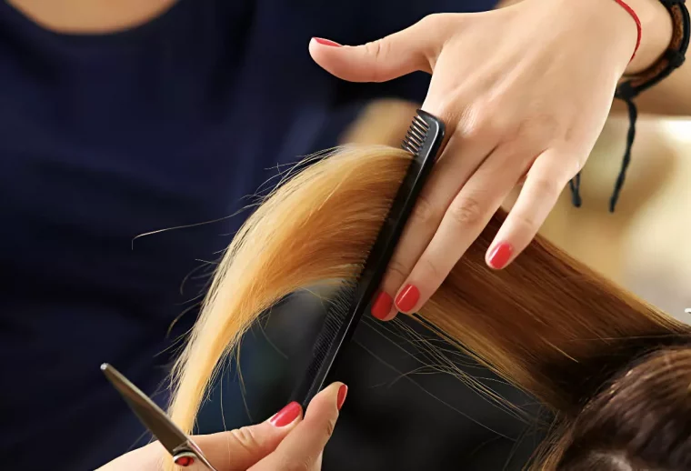 une main manicuree en rouge tient entre les doigts une meche de cheveux et passe le peigne avec la deuxieme main qui tient des ciseaux