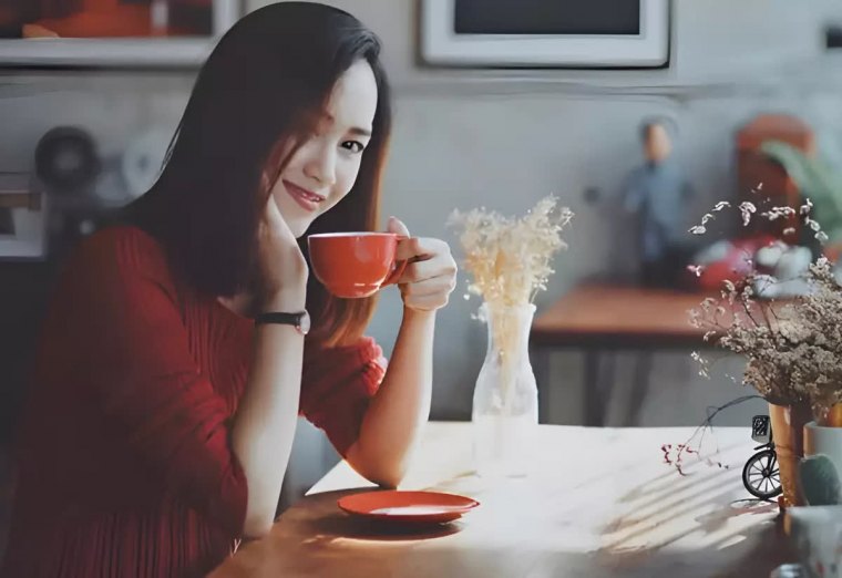femme asiatique assise sur une table porte une tasse rouge pres de son visage et sourit