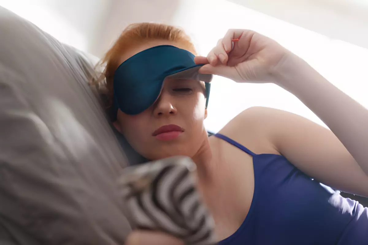 femme au lit qui regarde avec un œil sous son masque aux yeux l ecran de son portable