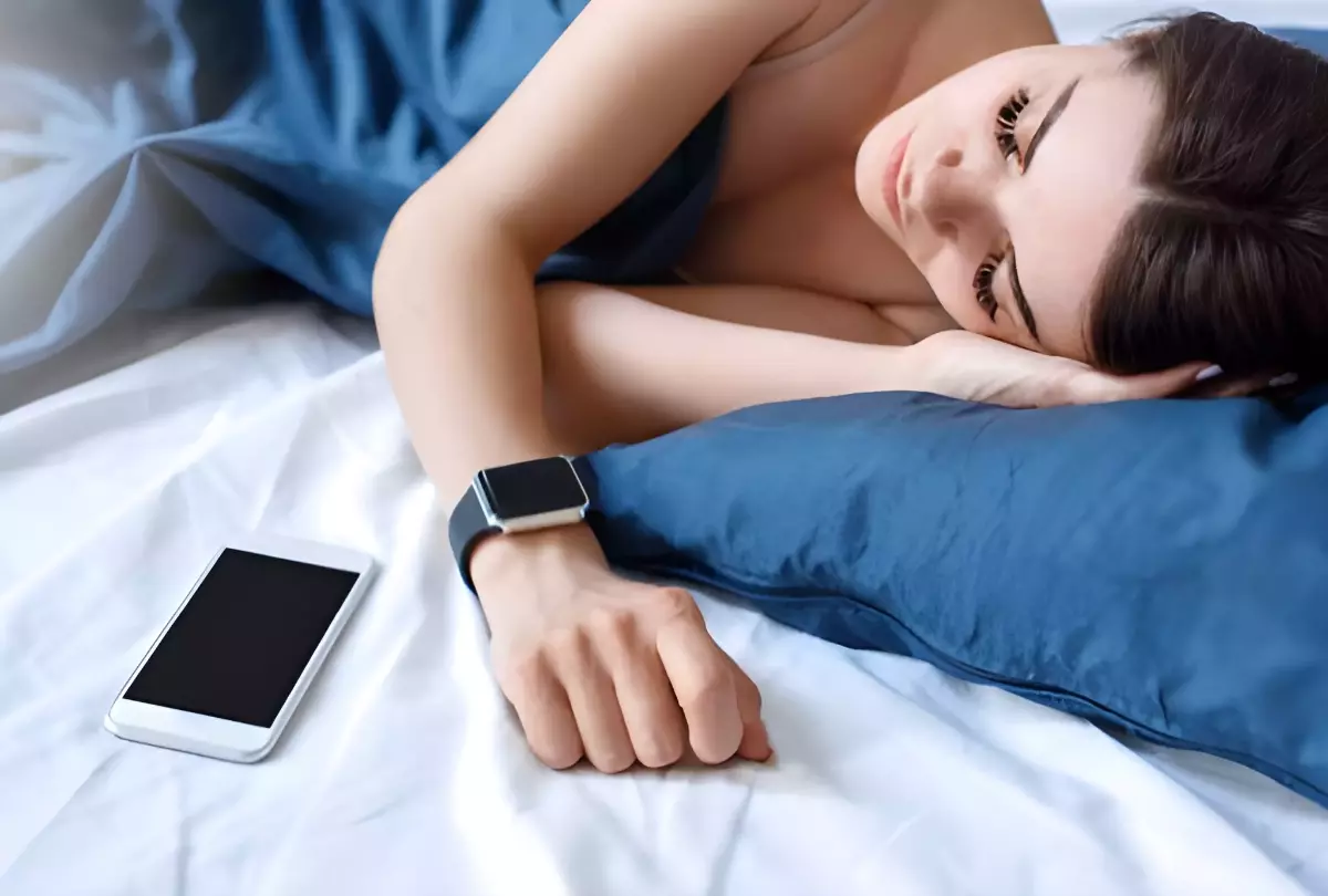 femme qui dort dans des draps blancs bleus avec une montre connectee et un telephone portable sur le lit a cote d elle