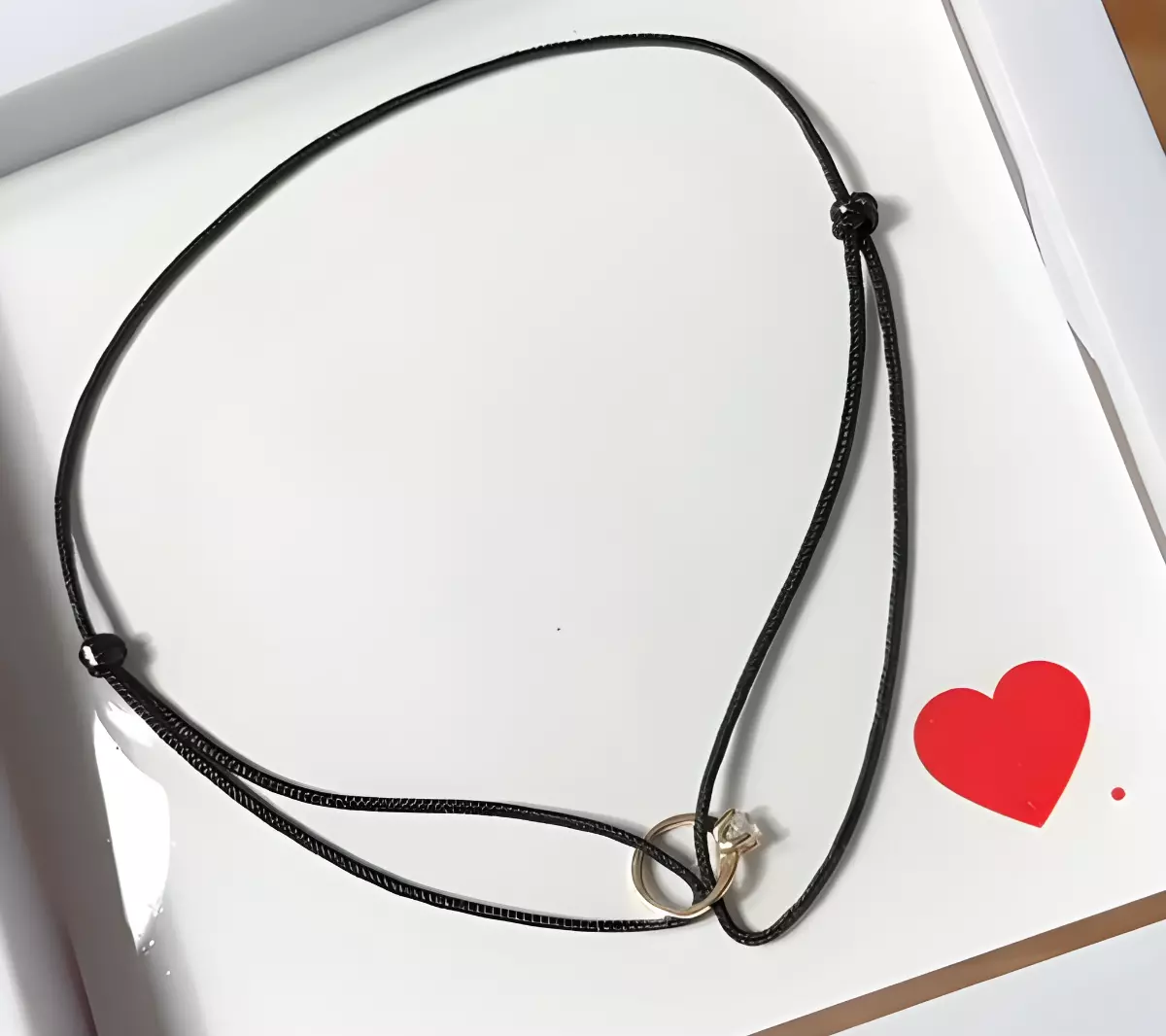 bracelet noir avec une mini bague de fiancailles avec une pierre dans une boite blanche et un petit coeur rouge en bas a droite
