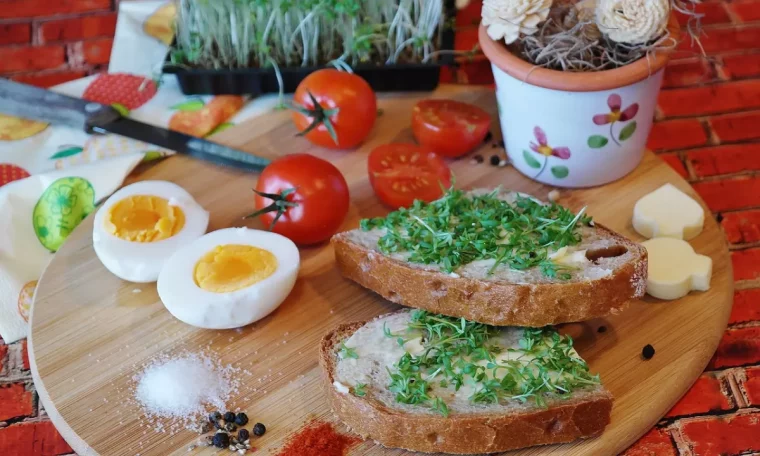 planche a decouper en bois recouverte de deux tartines un œuf cuit decoupe en deux tomates cerises epices fromage et un petit pot