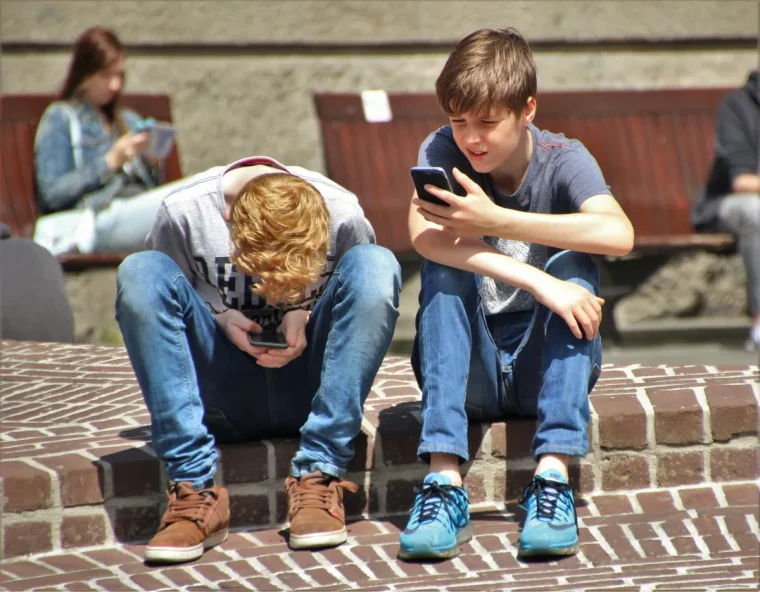 deux garcons assis sur le trotoir jeans bleus et t shirts jouent sur ces portables